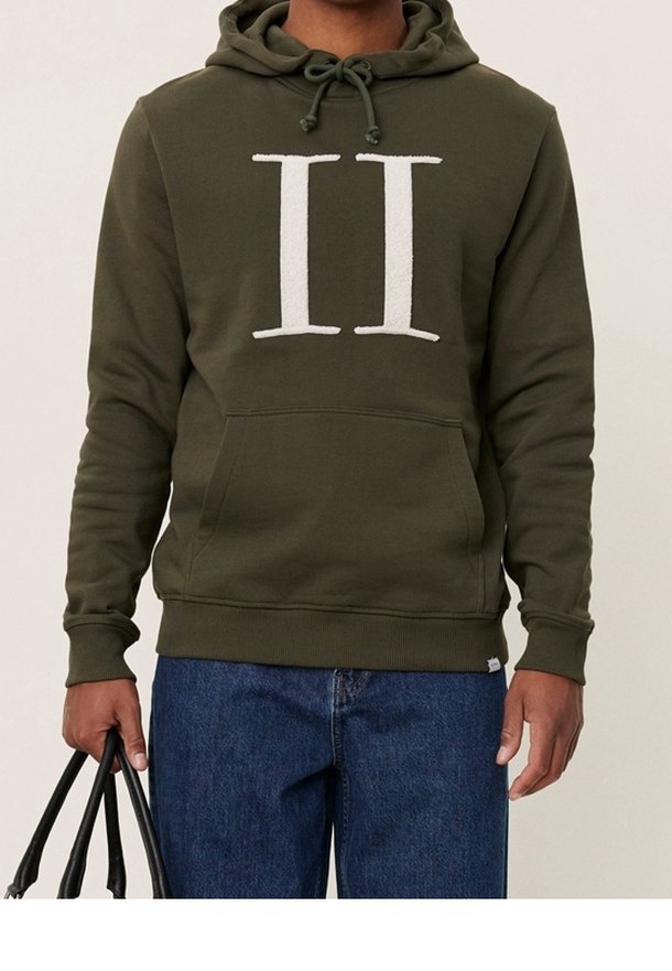 Olivgrön hoodie med en stor vit "II" emblem, dragsko-hoodie, frontficka och ribbade ärmslut. Bärs med blå denim och en svart väska.