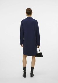 Mörkblå tröjdress med långa ärmar och hög krage. Har en avslappnad passform och når över knäna. Kombinerad med en liten svart handväska.