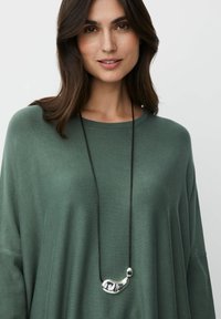 Grüner gestrickter Pullover mit weiten Ärmeln, kombiniert mit einer schwarzen Schnur-Halskette, die einen glatten, silbernen, abstrakten Anhänger zeigt.