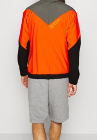 Man som bär en färgblockerat vindjacka i orange, svart och grått, med grå knälånga shorts, sedd bakifrån.