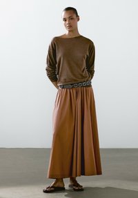 Hnědý svetr s dlouhými rukávy a širokým výstřihem, doplněný volnou sukní v barvě rezavé, která je držena černým opaskem s obláčkovými detaily.