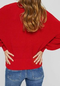 Roter gestrickter Pullover mit lockerer Passform, ausgestattet mit weiten Ärmeln und einem gerippten Saum. Kombiniert mit blauen Jeans.