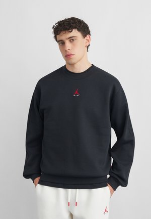 Młody mężczyzna z kręconymi włosami ubrany w czarną bluzę i białe spodnie dresowe, oba z czerwonym logo Jumpman.