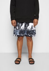 Sweatshirt noir et short de bain jusqu'aux genoux avec un motif abstrait noir, blanc et bleu. Des sandales noires complètent la tenue.