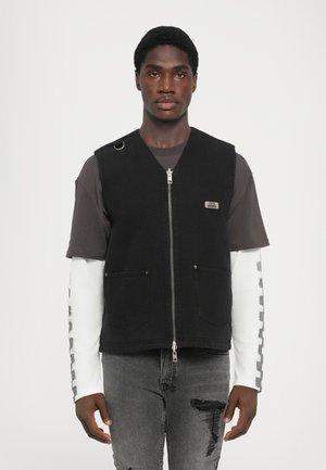 Sort denimvest med lynlåslukning, to forlommer og en ringdetalje. Bæres over en grå skjorte med hvide lange ærmer.