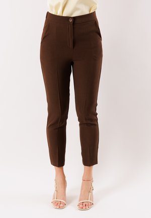 Pantaloni alla caviglia marroni slim-fit indossati con tacchi aperti beige, tagliati in vita, su uno sfondo chiaro e uniforme.