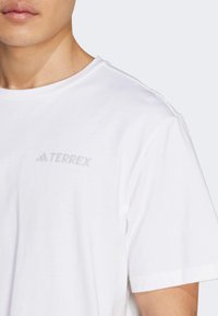 Biela bavlnená tričko s okrúhlym výstrihom a jemným šedým logom "TERREX" na hornej ľavej strane hrude. Krátke rukávy, hladká textúra.