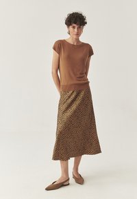 Frau mit kurzen lockigen Haaren, die ein braunes Kurzarmoberteil, einen mittelblauen Rock mit braun-schwarzen Flecken und tanfarbene Slingback-Sandalen trägt. Sie steht vor einem einfarbigen Hintergrund.