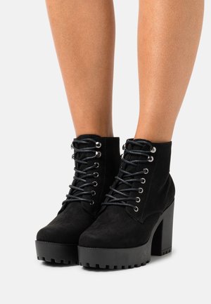 Black suede platform ankle boots with chunky heels and lace-up front worn on bare legs against white background.