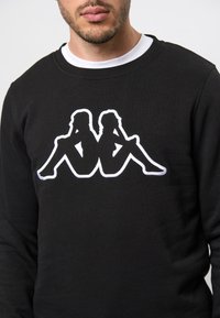 Zwarte sweatshirt met een witte omtrek graphic van twee zittende figuren. Het materiaal lijkt zacht en heeft een ronde hals.
