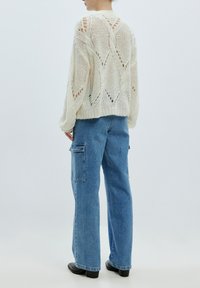 Strickpullover in Creme mit Lochmuster, lockerer Schnitt und geripptem Saum, kombiniert mit blauen, weit geschnittenen Jeans mit Seitentaschen.