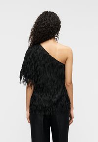 Μαύρο τοπ με έναν ώμο, διακοσμημένο με φράγκος, υφή υφάσματος και χαλαρή εφαρμογή. Η πίσω όψη παρουσιάζει ασύμμετρο σχέδιο και λεία άκρα.