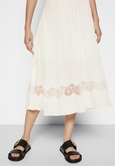 Robe blanche plissée avec un ourlet en dentelle festonnée de motif floral, associée à des sandales noires avec une boucle en or.