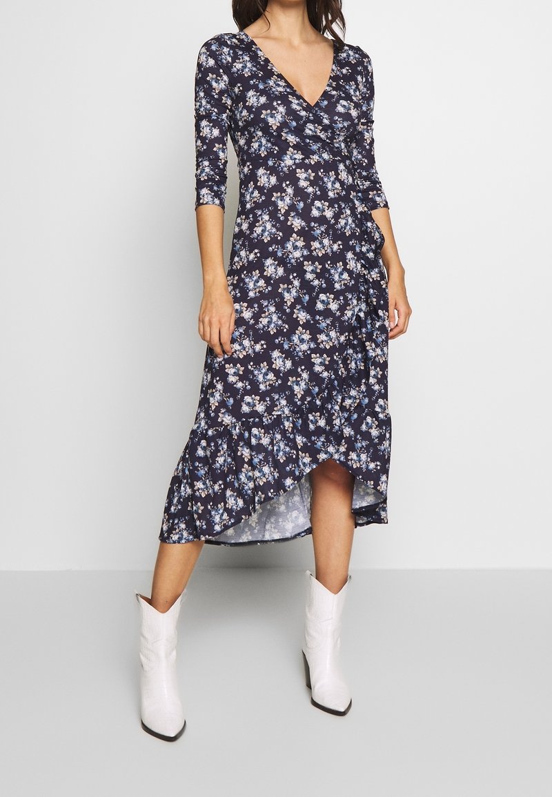 Robe portefeuille florale en bleu marine avec des manches trois-quarts, ornée de fleurs bleues et blanches, à ourlet volanté, accompagnée de bottes de cowboy blanches.
