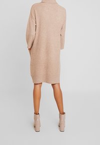 Abito maglione beige con collo alto, vestibilità ampia e maniche a tre quarti; abbinato a stivali alla caviglia di colore chiaro.