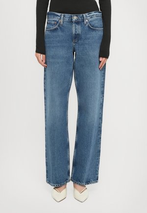Personne portant un jean en denim bleu à jambe droite, un haut noir à manches longues et des chaussures blanches à bout pointu sur un fond gris clair.