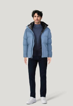 Light blue puffer jacket with a black collar, paired with a navy sweater and dark pants. Model wears white sneakers, standing against a gray backdrop.