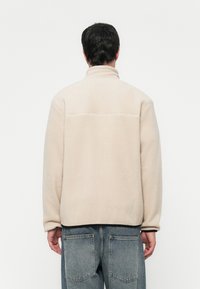 Beige fleecejakke med høy krage, litt oversized passform og teksturert overflate. Svart kant langs bunnen og ermene.