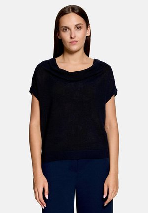 Femme aux cheveux bruns raides portant un haut noir ample à manches courtes et un pantalon foncé sur un fond clair uni.