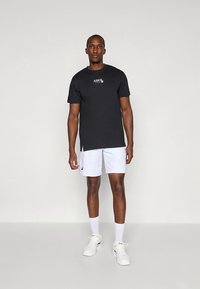 Black short-sleeve t-shirt with a small colored logo, paired with white athletic shorts, and white sneakers, on a neutral background.