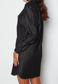 Svart denimklänning med knäppning framtill, långa ärmar med knappmanchetter och rundad fåll. Texturerat tyg med en svag lyster.