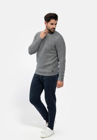 Grauer Strickpullover mit gerippten Bündchen, kombiniert mit dunkelblauen Jogginghosen. Weiße Sneaker runden das Outfit ab. Einfarbiges Hintergrund.