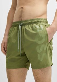 Green swim shorts made of lightweight, textured fabric, featuring an elastic waistband, drawstring, and a large brand logo on the side.