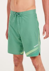 Groene zwembroek met een mesh-paneelontwerp aan de zijkanten, een koord in de taille en het woord "PROTEST" in het wit gedrukt.