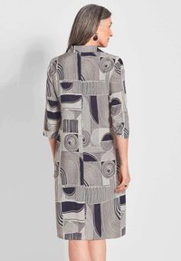 Langmouw jurk in beige met donkere geometrische patronen, met cirkelvormige en lineaire ontwerpen. Stof lijkt glad; knie-lange snit.