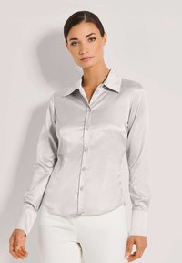 Frau trägt eine hellgraue satinierte Bluse mit Knöpfen und langen Ärmeln sowie weiße Hosen, steht vor einem schlichten hellen Hintergrund.