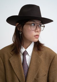 Brown felt hat, clear glasses, brown wool blazer, white shirt, blue tie with orange pattern. Hair styled down, subtle silver earring.