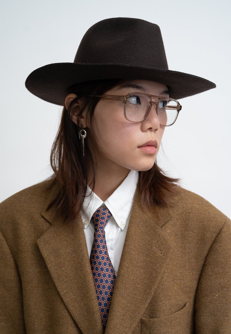 Brown felt hat, clear glasses, brown wool blazer, white shirt, blue tie with orange pattern. Hair styled down, subtle silver earring.