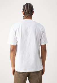 Pessoa com cabelo trançado, vestindo uma t-shirt branca simples de mangas curtas e calças castanhas, de pé de costas contra um fundo liso.