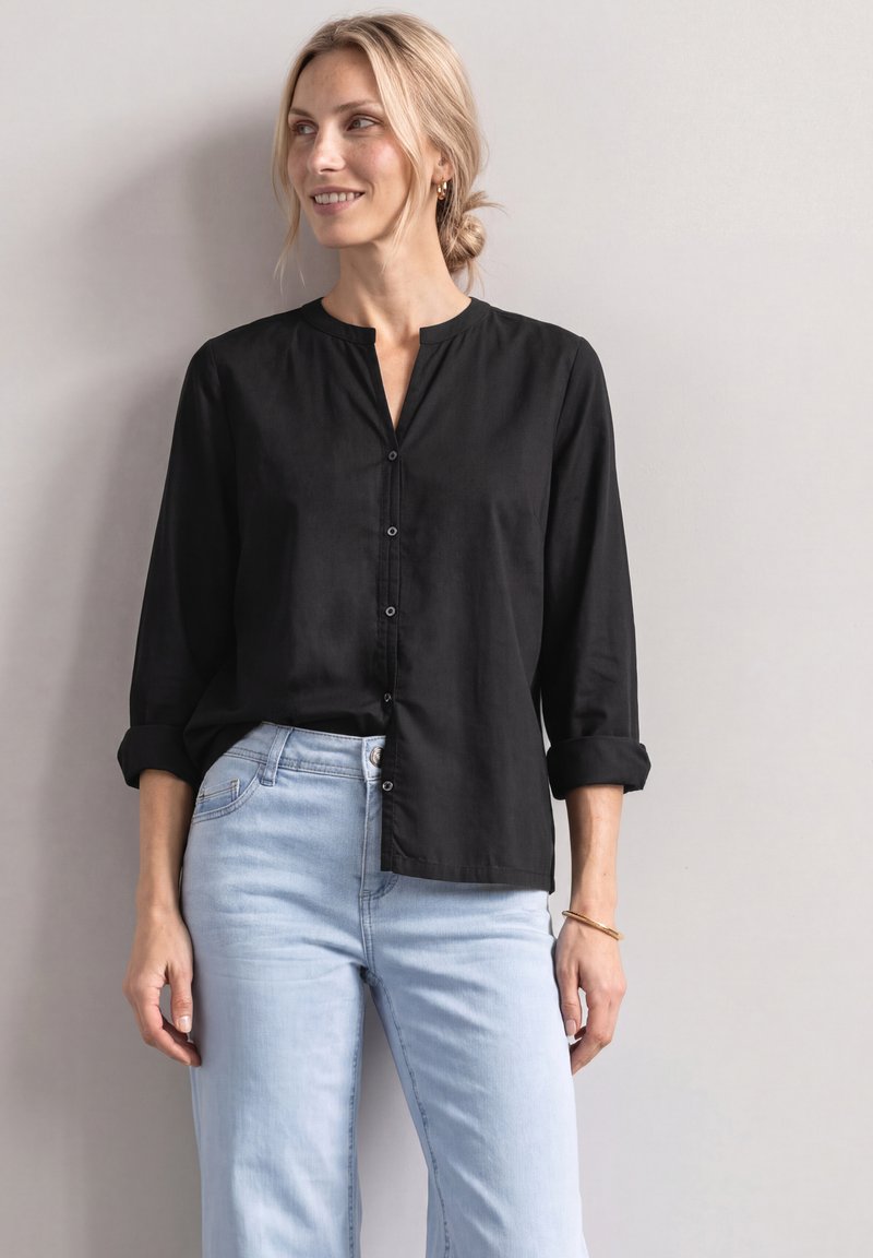 Femme aux cheveux blonds en chignon bas, portant un chemisier noir boutonné et un jean bleu clair, debout contre un mur clair uni, souriante et regardant vers la droite.