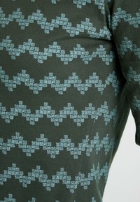 Zielony materiał z wzorem w zygzaki w jasnoniebieskim. Tekstura jest gładka, a geometryczne kształty tworzą powtarzający się wzór na materiale.