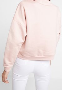 Vrouw die een lichtroze sweatshirt met lange mouwen en witte strakke jeans draagt, weergegeven van achteren, staand tegen een effen witte achtergrond.