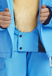Blauwe winterjas met bontvoering, gekenmerkt door zwarte drukknoopsluitingen, elastische manchetten en een gladde buitenkant. Gedetailleerde stiksels zichtbaar.