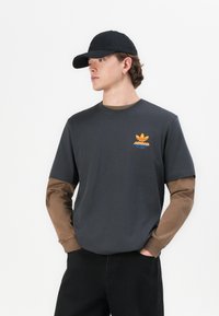 Szürke, rövid ujjú póló barna hosszú ujjal, narancssárga és kék Adidas logóval a mellkason. Fekete baseball sapka egészíti ki a megjelenést.