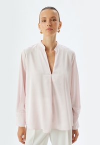 Blusa a maniche lunghe di colore rosa in un tessuto liscio con scollatura a V e dettagli di cucitura sottili. Presenta polsini aderenti e una vestibilità rilassata.