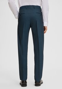 Man in op maat gemaakte donkerblauwe pantalon, witte shirt en zwarte dresschoenen, staand tegen een effen witte achtergrond, rugaanzicht.