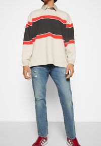 Hombre vestido con un polo de mangas largas a rayas beige, negro y rojo, jeans azules rasgados, y zapatillas rojas y blancas, de pie contra un fondo blanco.