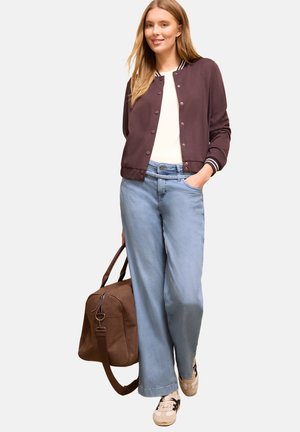 Ung kvinde iført en brun knappet jakke, hvid top, lyseblå vide jeans og sneakers, der holder en stor brun rejsetaske.