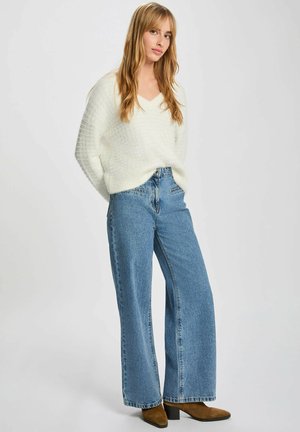 Giovane donna con capelli lunghi che indossa un maglione bianco peloso, jeans blu a gamba larga e stivali con tacco marroni, in piedi su uno sfondo neutro.