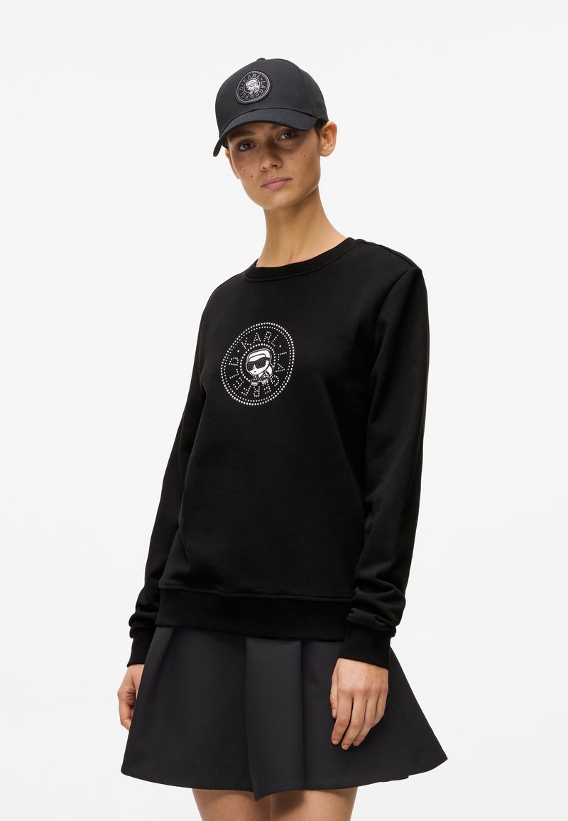 Γυναίκα με μαύρο φούτερ μακρυμάνικο με κυκλικό λογότυπο με τρουκς και καπέλο σε μαύρο χρώμα, συνδυασμένα με μαύρη πλισέ φούστα.