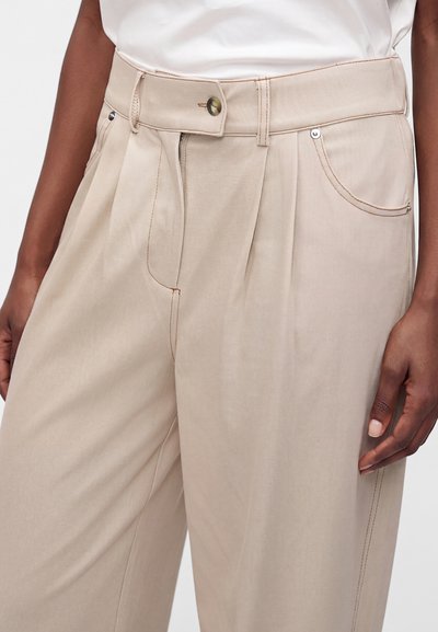 Gros plan sur un pantalon beige taille haute avec fermeture par bouton et zip, poches latérales, et un top blanc rentré, porté par une personne à la peau mate.