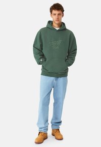 Grön hoodie med texten "Ärlighet Råd" , frampå ficka, bärs med ljusblå jeans och bruna kängor. Avslappnad passform, mellantjockt tyg.