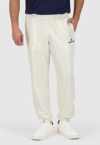 Crème kleurige trainingsbroek met een elastische tailleband, taps toelopende boorden en een logo op de linkerdij. Draagt met witte sneakers.