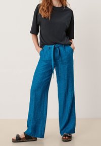 Pantalon large en lin bleu avec taille nouée, associé à un t-shirt noir à manches courtes et des sandales plates noires. Texture naturelle visible.