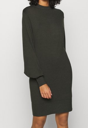 Femme portant une robe en maille vert foncé à manches longues avec poignets et ourlet côtelés, debout devant un fond uni gris clair.