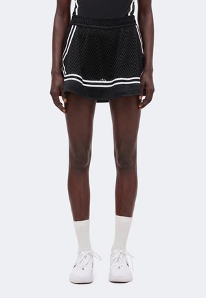 Personne portant une jupe de sport en filet noire avec des bandes blanches, des chaussettes blanches montantes et des baskets blanches, debout devant un fond clair uni.